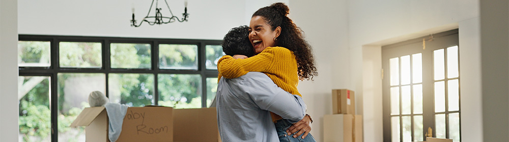A young couple hugs in excitement while moving boxes are stacked in the background.